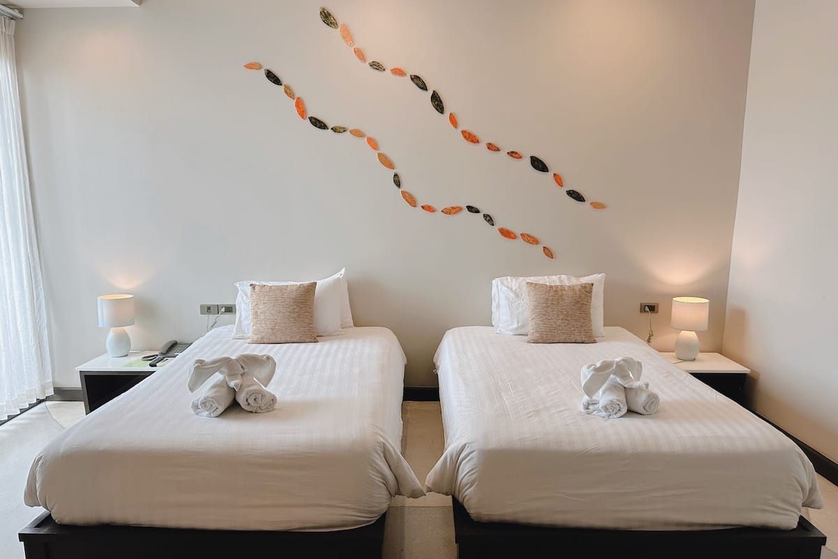 A hotel room with two beds, each with a pillow and a towel. There are two bedside tables with lamps.