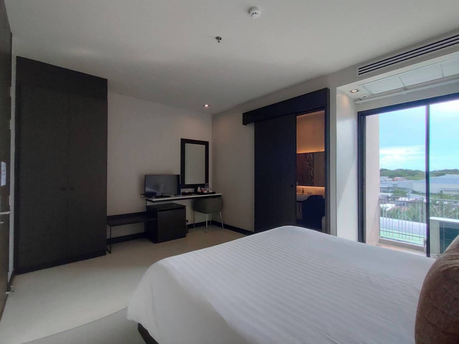 A modern hotel room with a large bed, a desk with a computer, a mirror, and a view of the city through a large window.