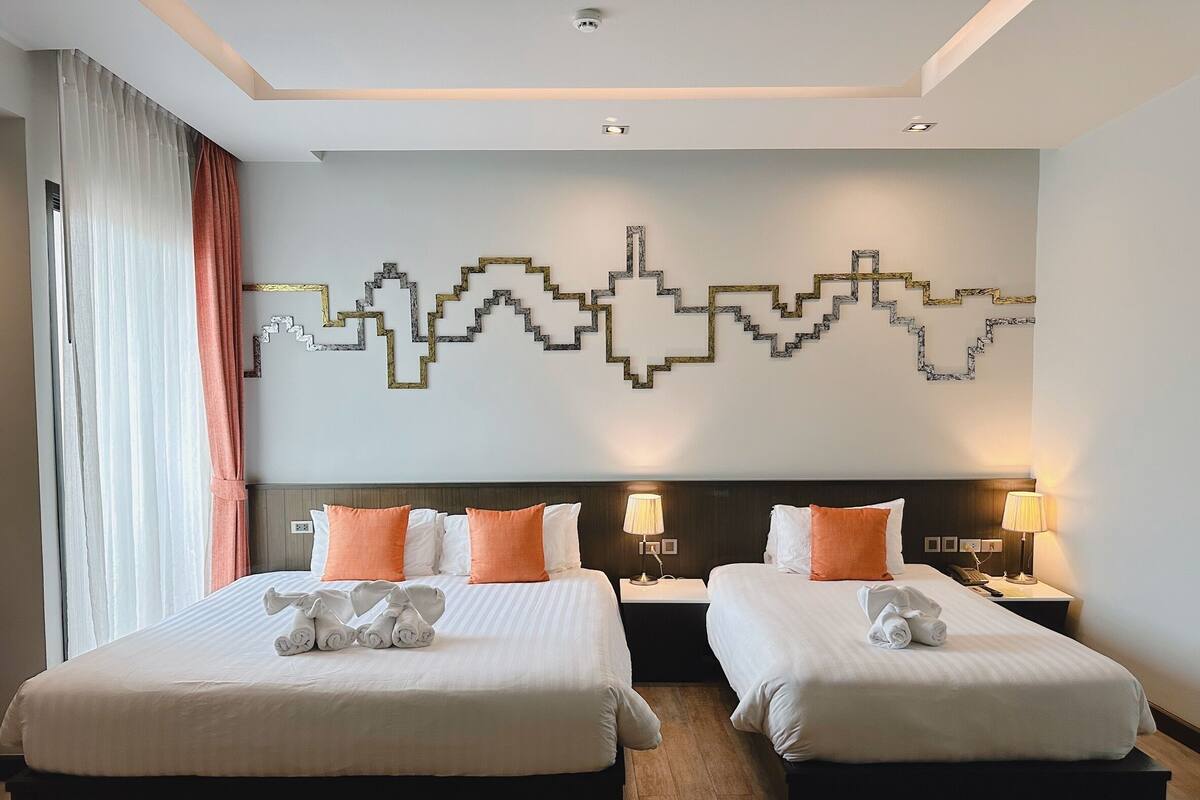 A hotel room with two beds, each with white linens and orange pillows, a modern wall art piece, and a wooden floor.