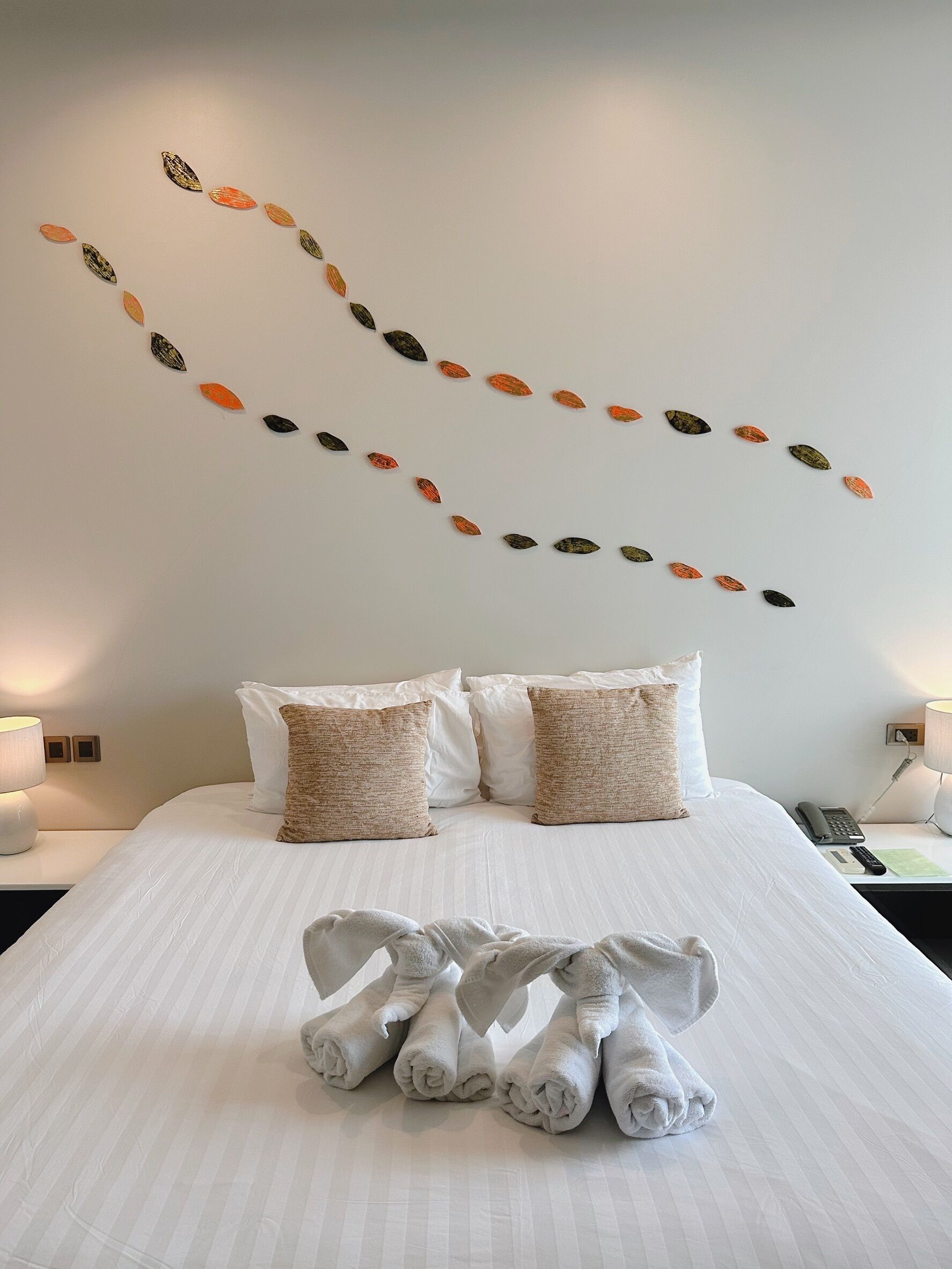 A neatly made bed with white linens and two beige pillows, a wall with a leaf pattern, and a bedside table with a lamp and a phone.