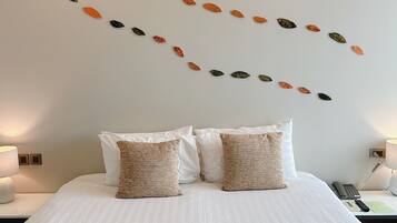 A neatly made bed with white linens and two beige pillows, a wall with a leaf pattern, and a bedside table with a lamp and a phone.