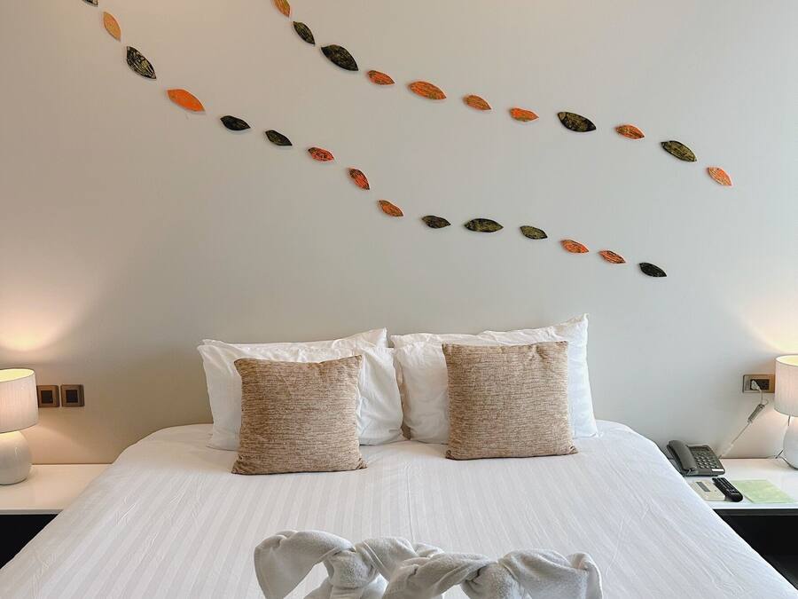 A neatly made bed with white linens and two beige pillows, a wall with a leaf pattern, and a bedside table with a lamp and a phone.