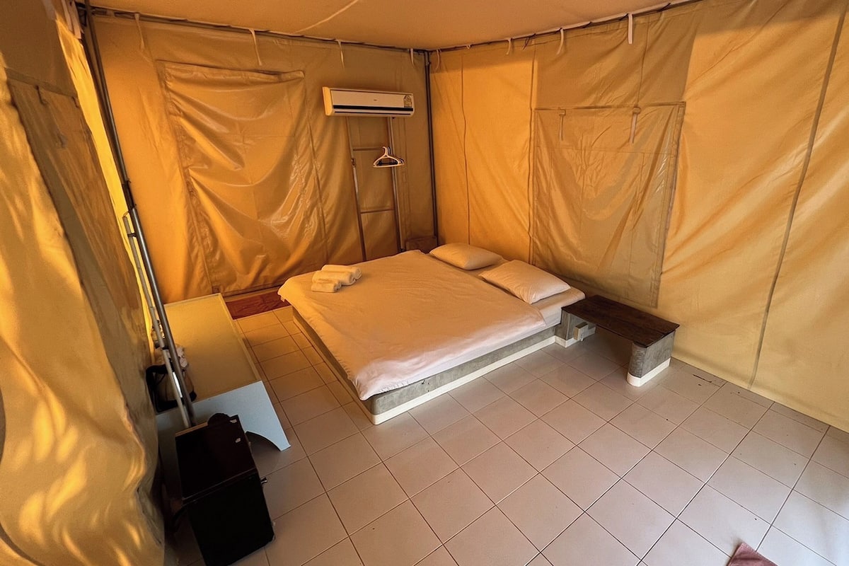 A single bed with a mattress and pillows, a bedside table with a lamp, a hanging clothes rack, and a small bench.