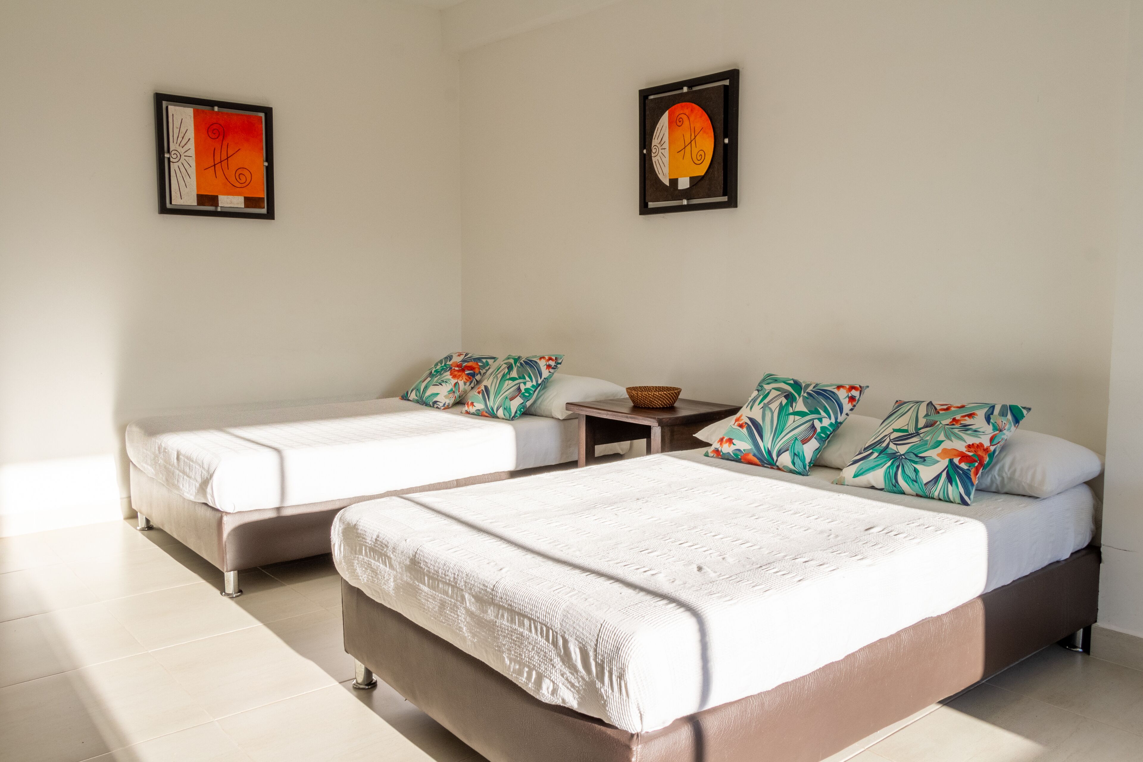 A hotel room with two beds, a small table, and two framed artworks on the walls.