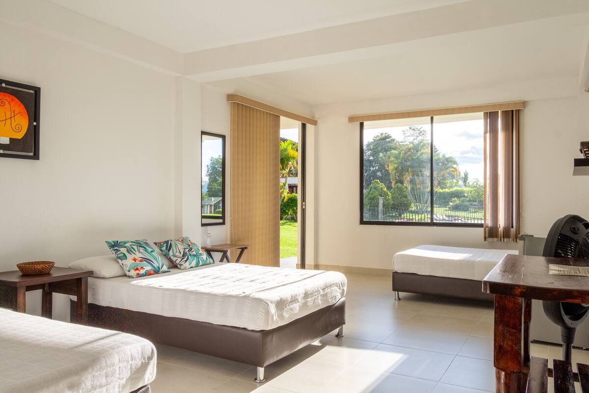 A spacious bedroom with a large bed, a smaller bed, and a desk. There is a window with a view of greenery outside.