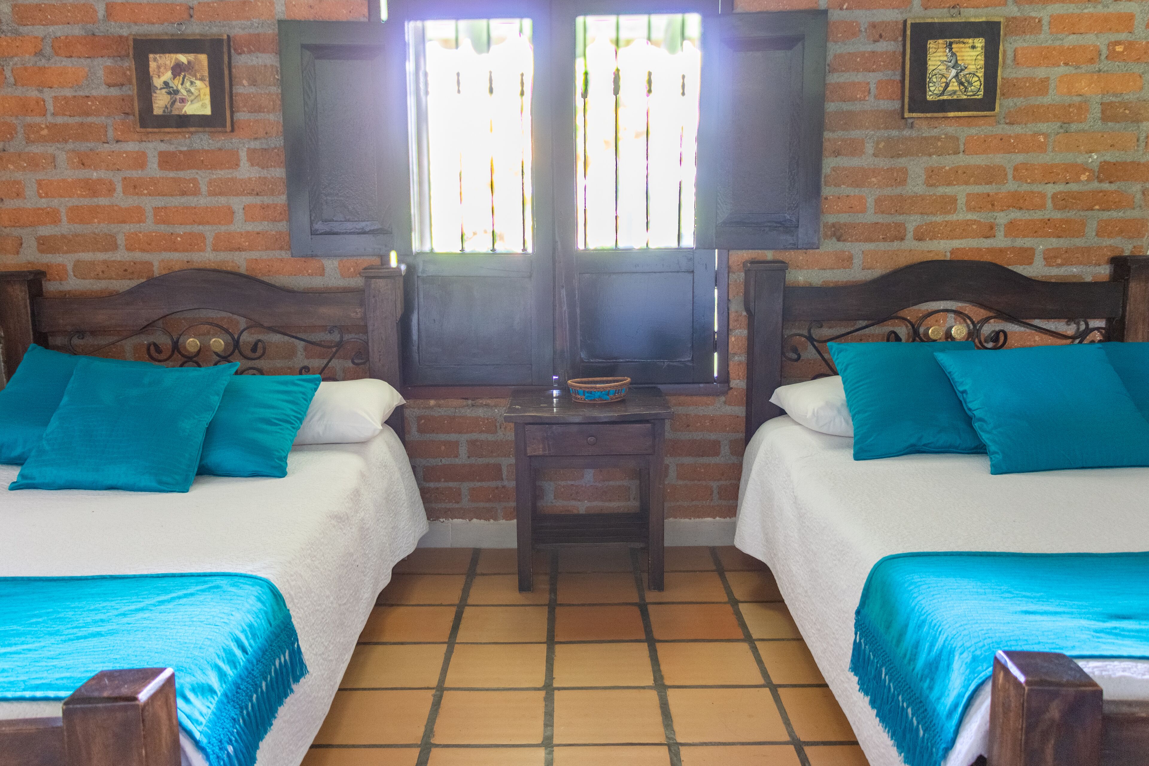 Habitación con dos camas, cada una con cabecero de madera, una mesita pequeña y un ventanal con contraventanas.
