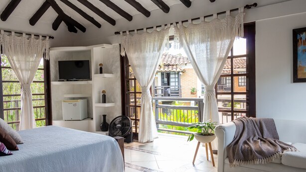 A bedroom with a bed, a TV, a fan, and a balcony with a view of greenery.