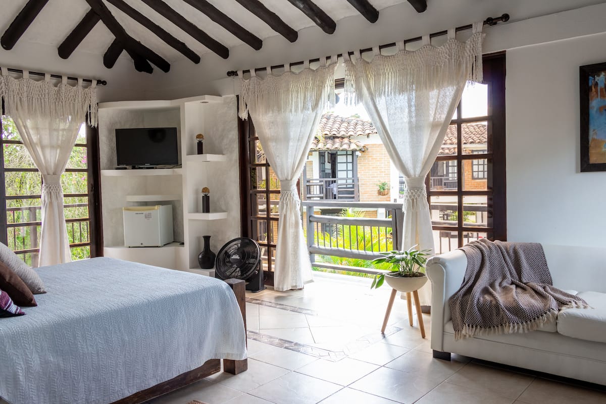 A bedroom with a bed, a TV, a fan, and a balcony with a view of greenery.