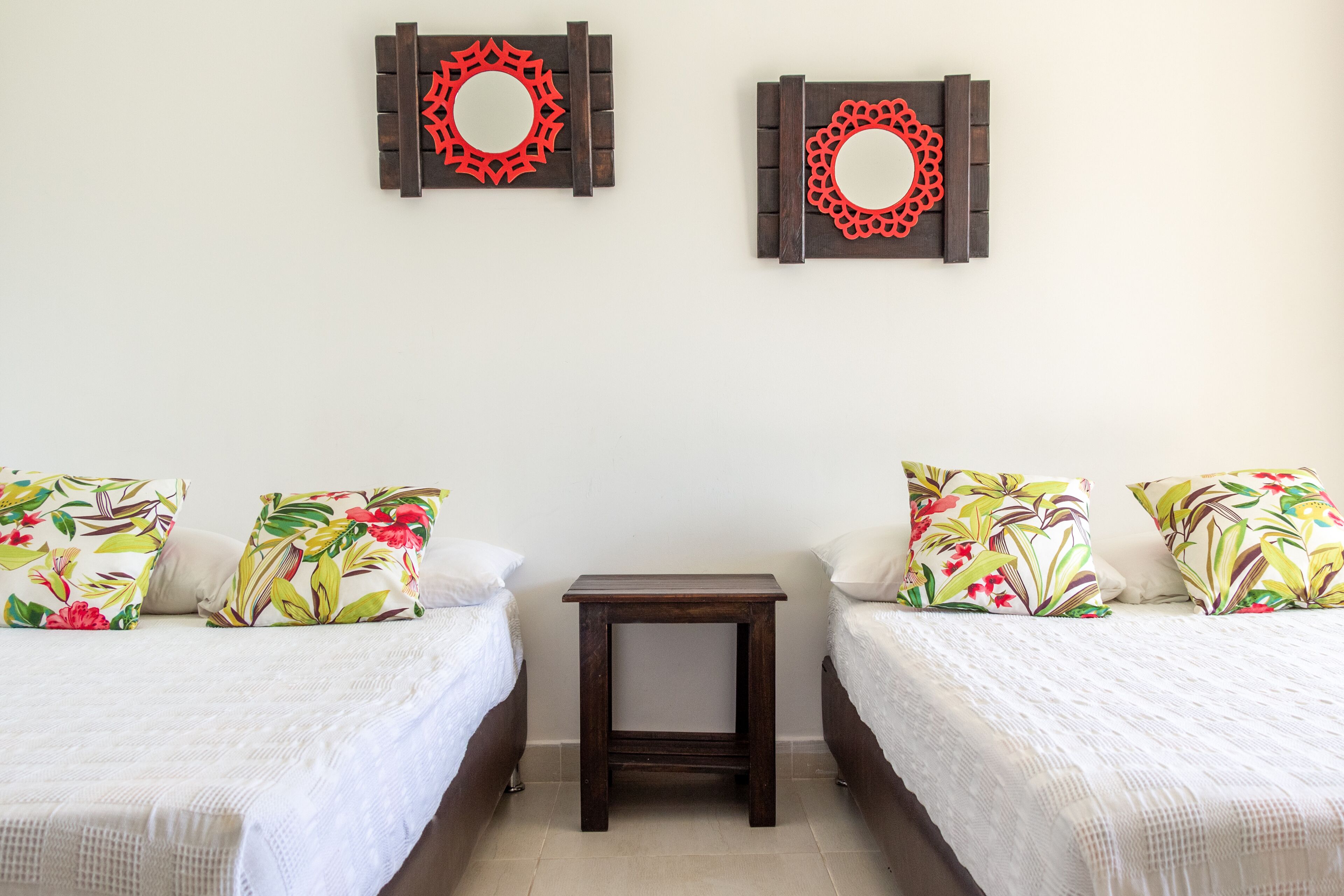A bedroom with two beds, each with floral-patterned pillows, a small wooden nightstand, and two wall-mounted decorative mirrors.