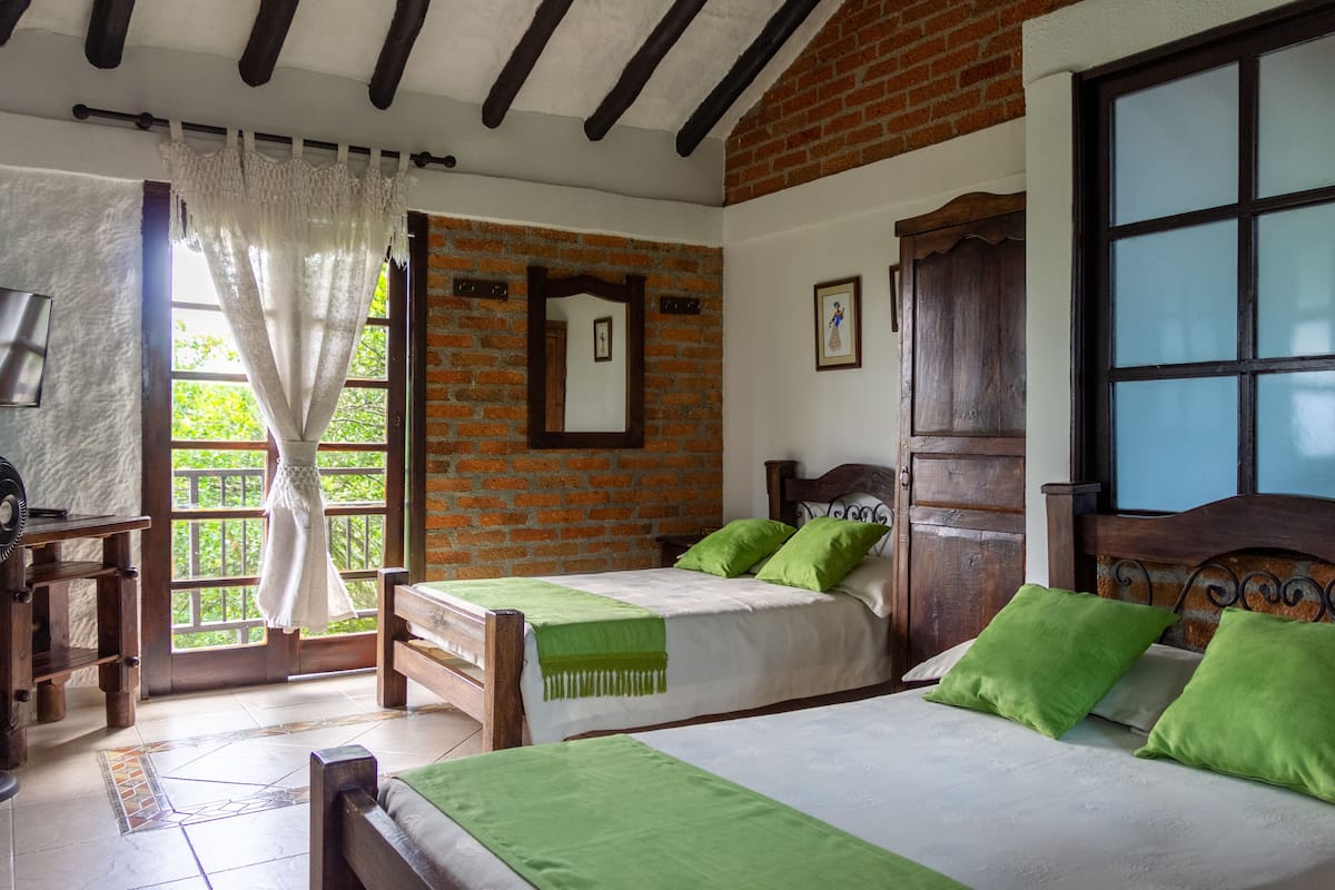 A room with two beds, a wooden desk, a mirror, and a window with a view of greenery.
