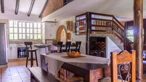 A dining area with a brick wall, wooden table, and chairs. There is a staircase with a railing leading to an upper level.