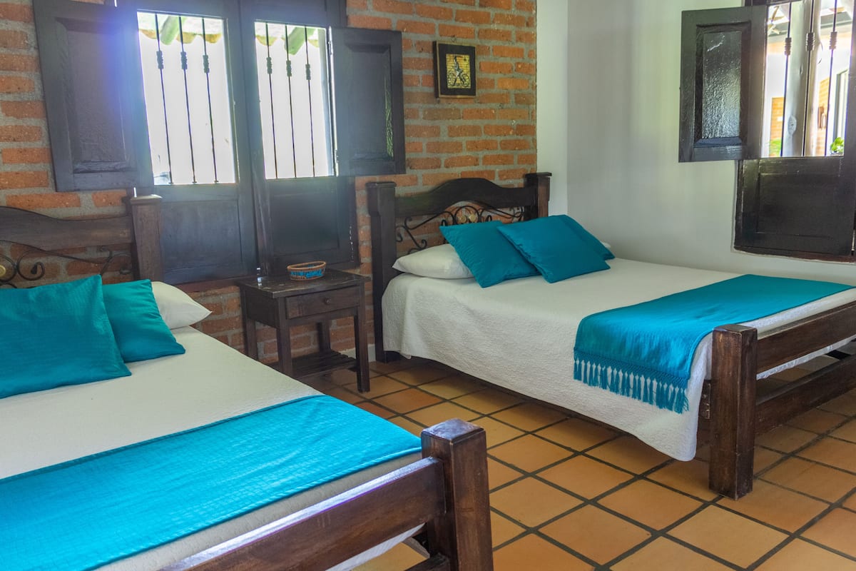 Two single beds with wooden frames and teal bedding, a small table with a bowl, and a brick wall with a window and a mirror.