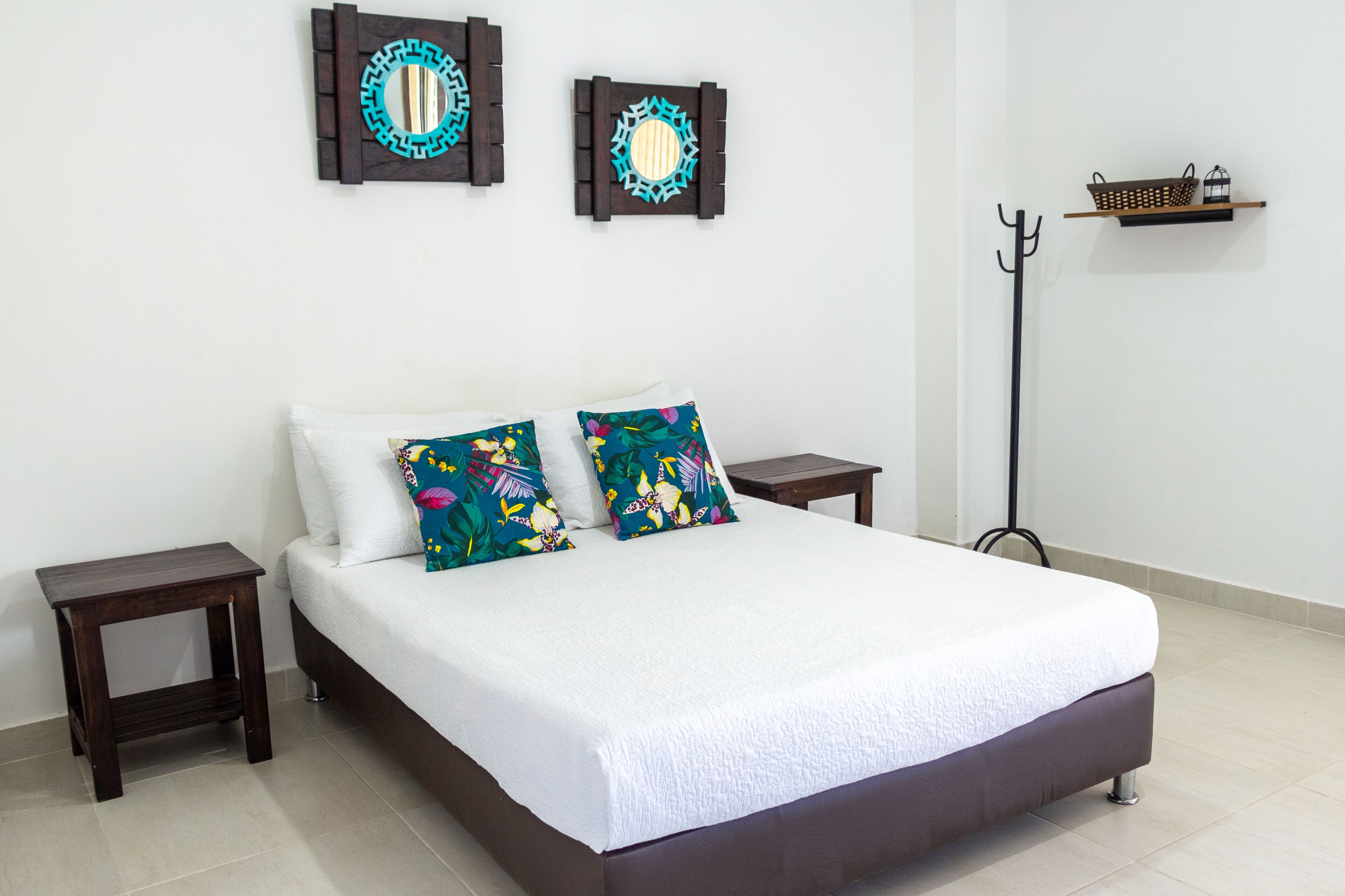 A neatly made bed with white bedding and colorful pillows, two wall-mounted mirrors, a small wooden shelf, and a floor lamp.