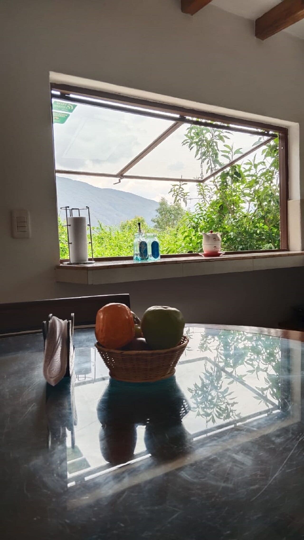 A room with a large window, a table with a basket of fruit, and a hanging plant.
