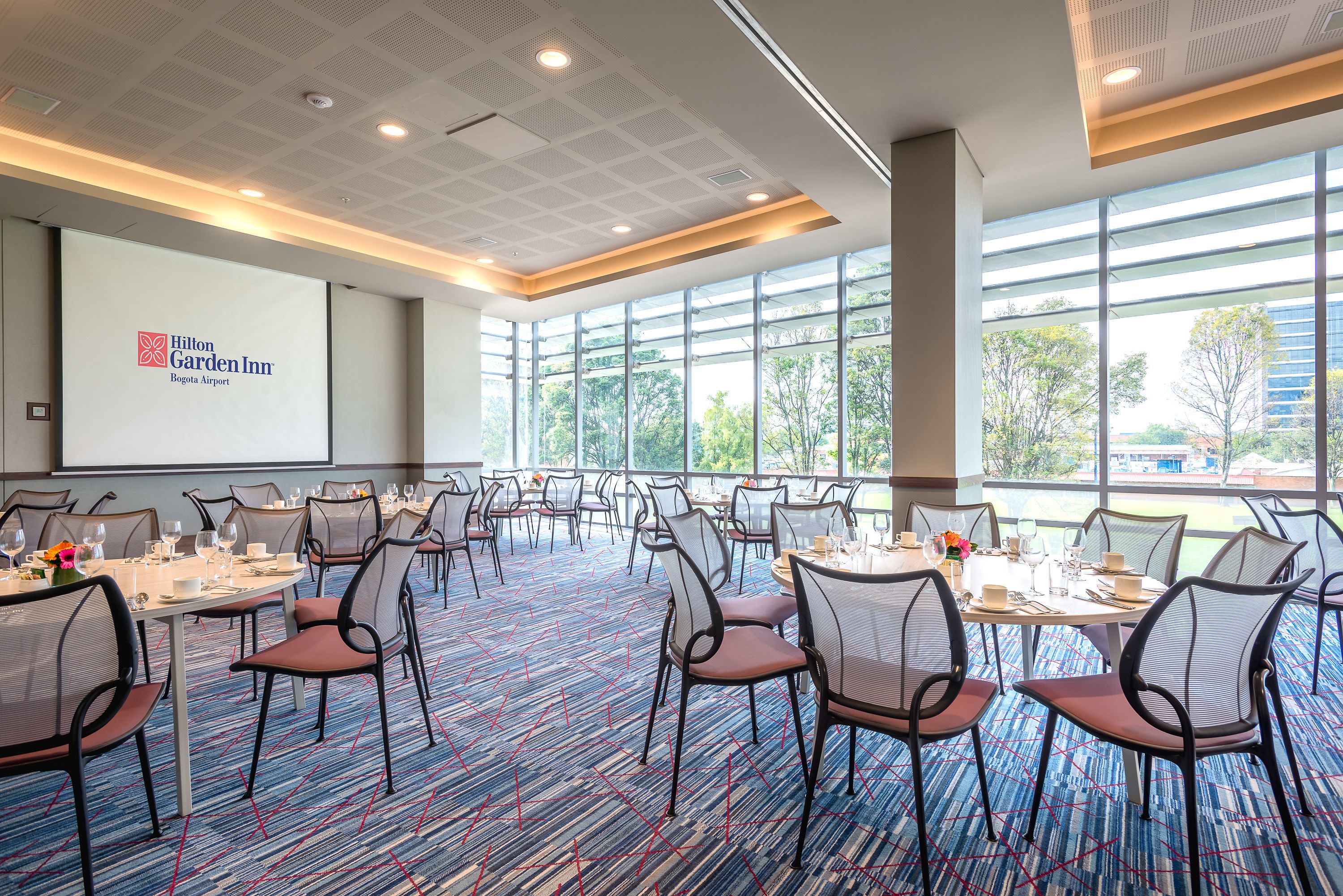 A conference room with tables set for a meeting, a large screen displaying the Hilton Garden Inn logo, and large windows offering a view of the outdoors.