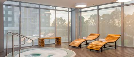 A modern indoor pool area with a hot tub, wooden loungers, and large windows offering views of the city.