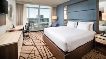 Habitación de hotel con una cama grande, un escritorio, una silla, un televisor y vistas a la ciudad a través de amplias ventanas.