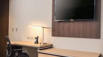 A hotel room with a wooden desk, a flat-screen TV mounted on the wall, a desk lamp, a chair, and a carpeted floor.