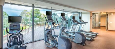 Un gimnasio con cintas de correr, elíptica y bicicleta estática, amplias ventanas con vistas a árboles y edificios, y un suelo pulido.
