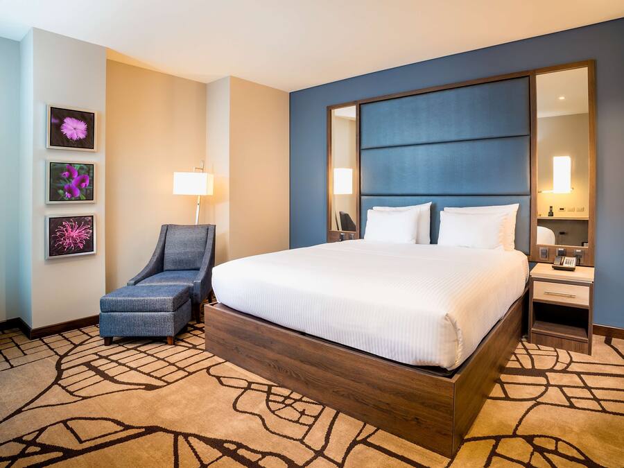 A hotel room with a large bed, a chair, a desk with a phone, and framed pictures on the wall.