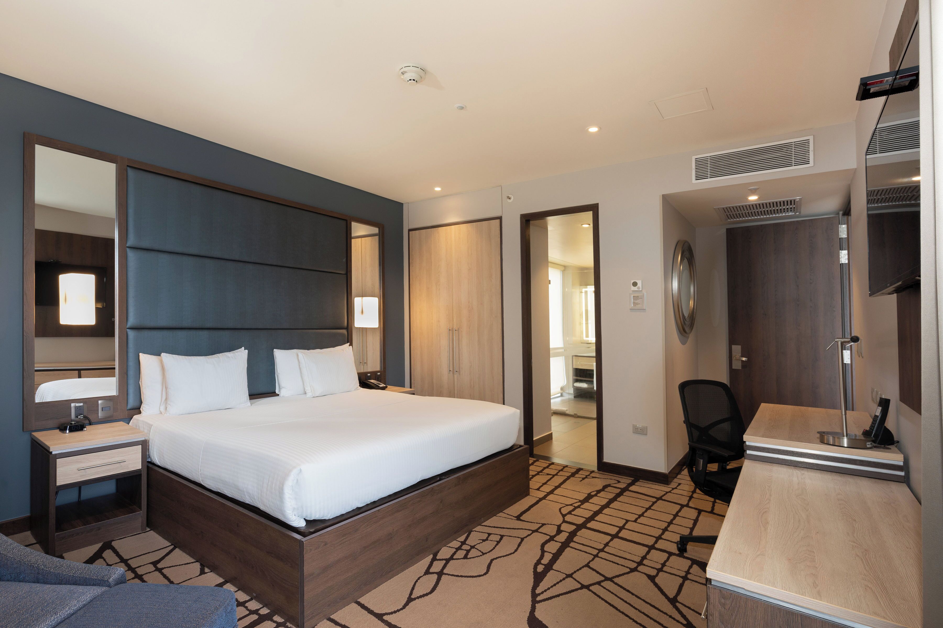 Una habitación de hotel moderna con una cama grande, mesitas de noche, un escritorio y un espejo.