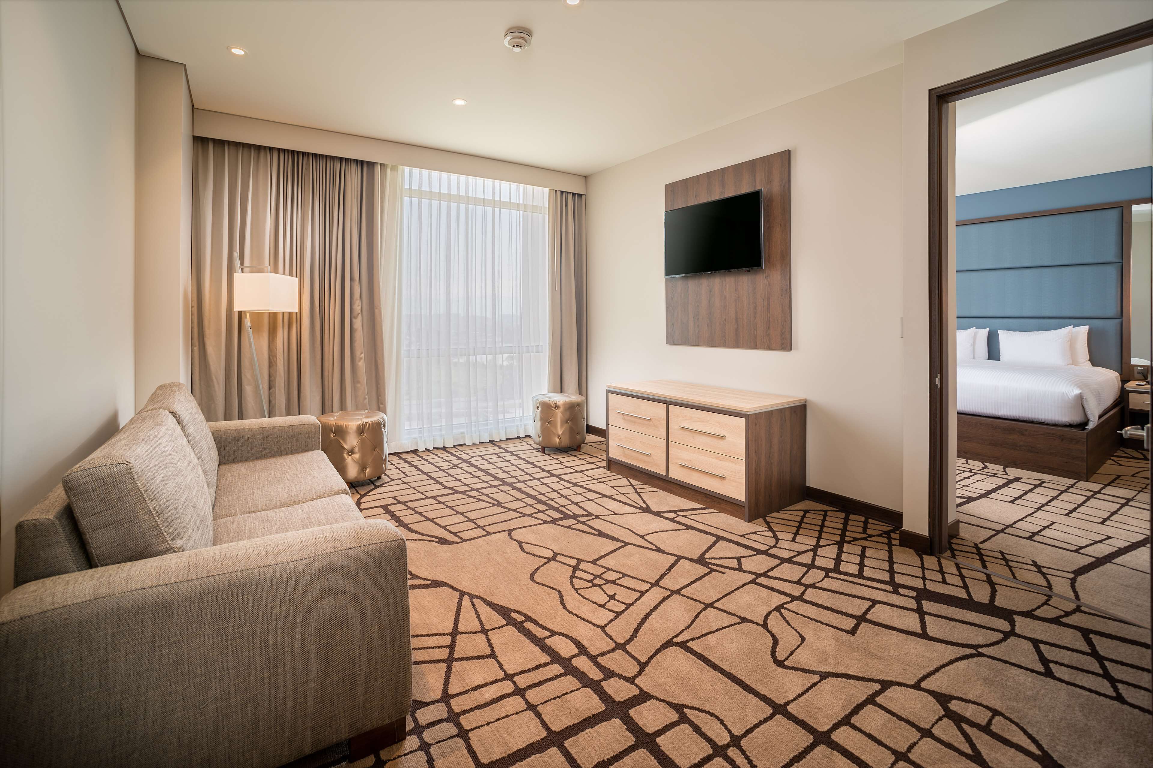 Une chambre d’hôtel avec un canapé, un téléviseur fixé au mur, un lit avec une tête de lit et un grand miroir.