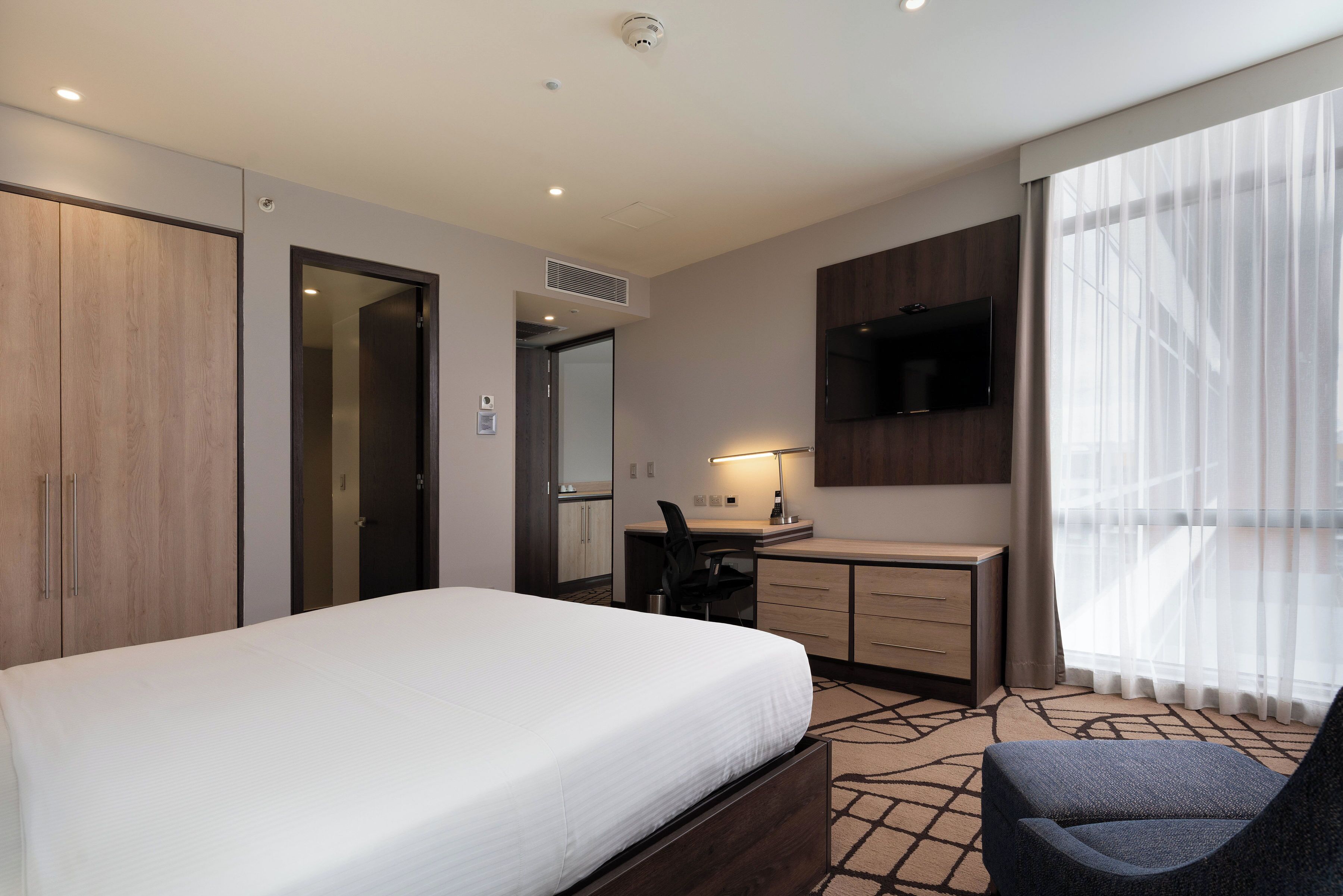 A hotel room with a bed, desk, chair, TV, and large windows.