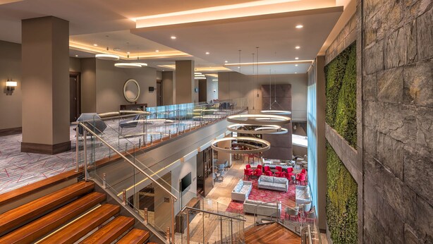 Un lobby moderno con una escalera de cristal, un espacio de descanso con sillas rojas y una pared verde.