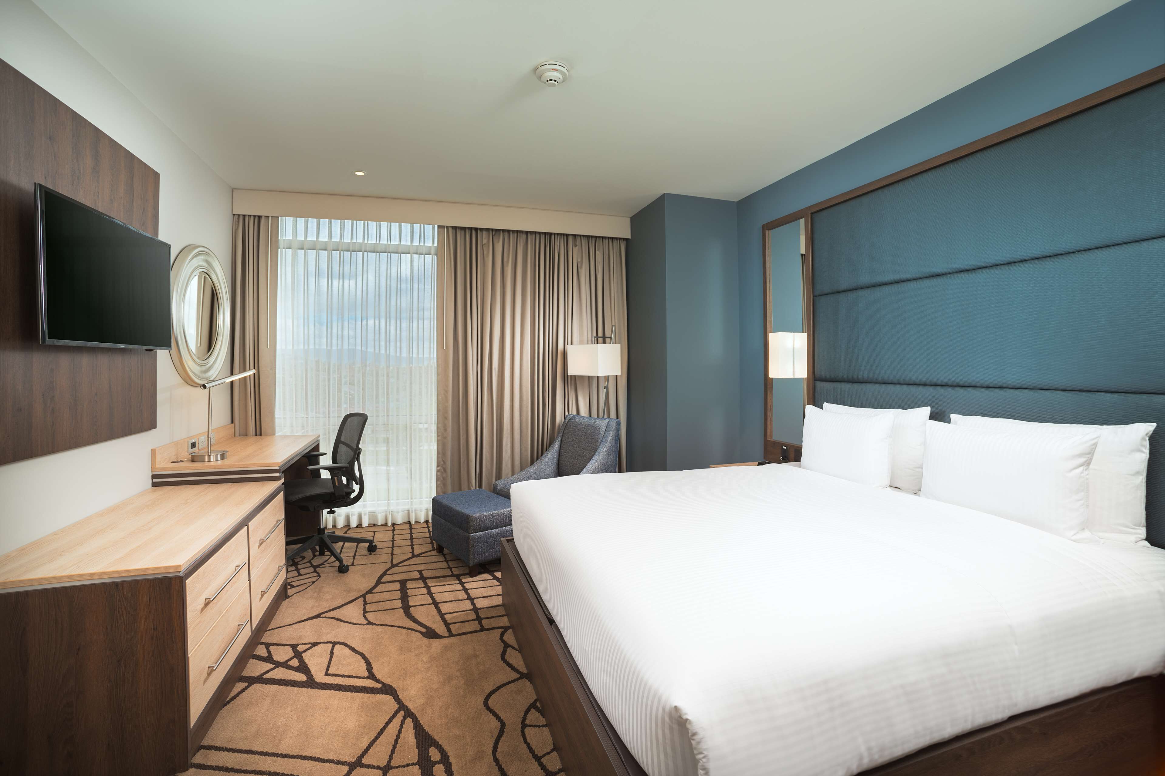 Habitación de hotel con una cama grande, un escritorio con silla, televisor montado en la pared y una ventana con cortinas.
