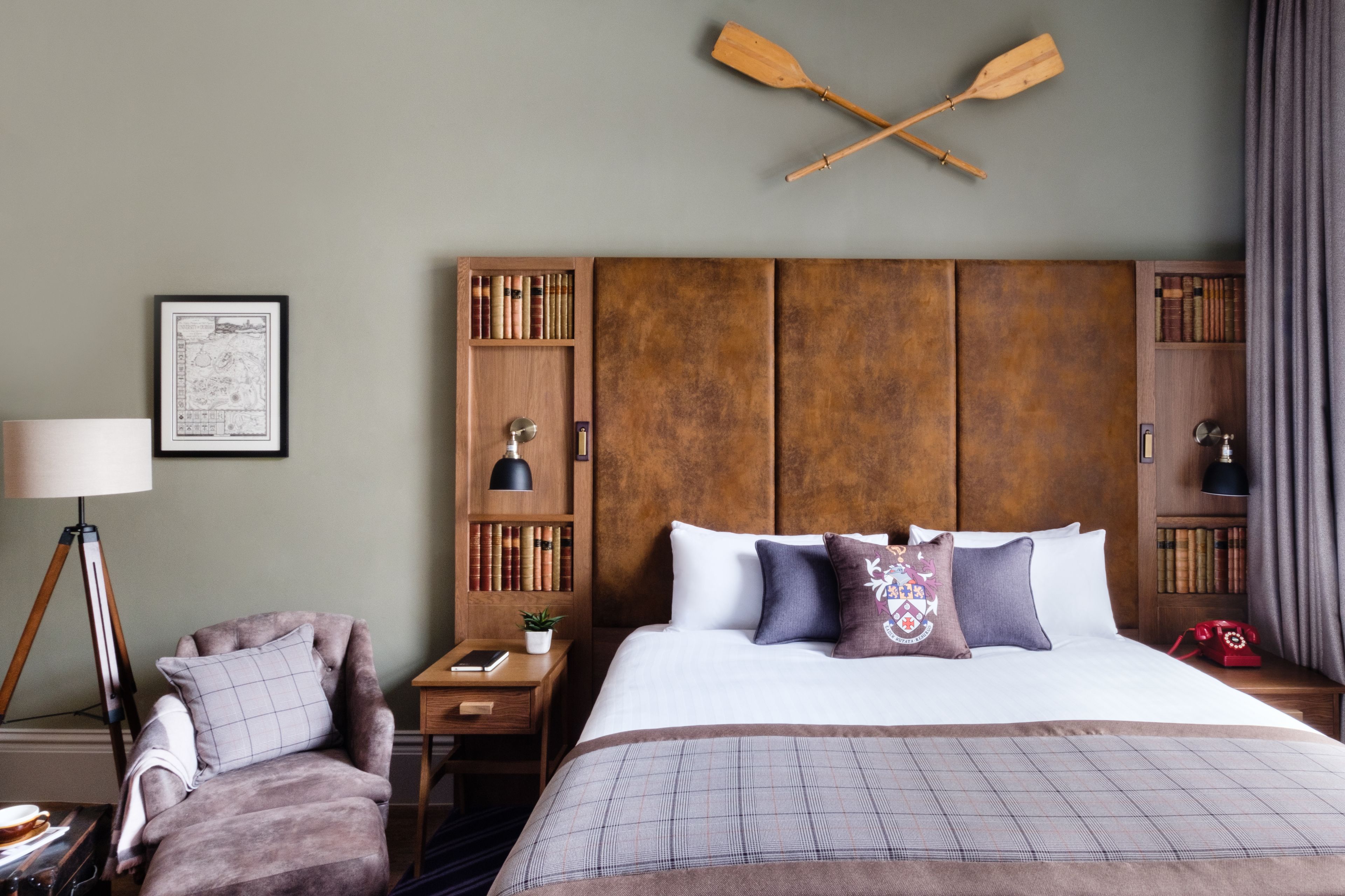 A hotel room with a large bed, a wooden headboard, a desk with a lamp, a chair, and a bookshelf.