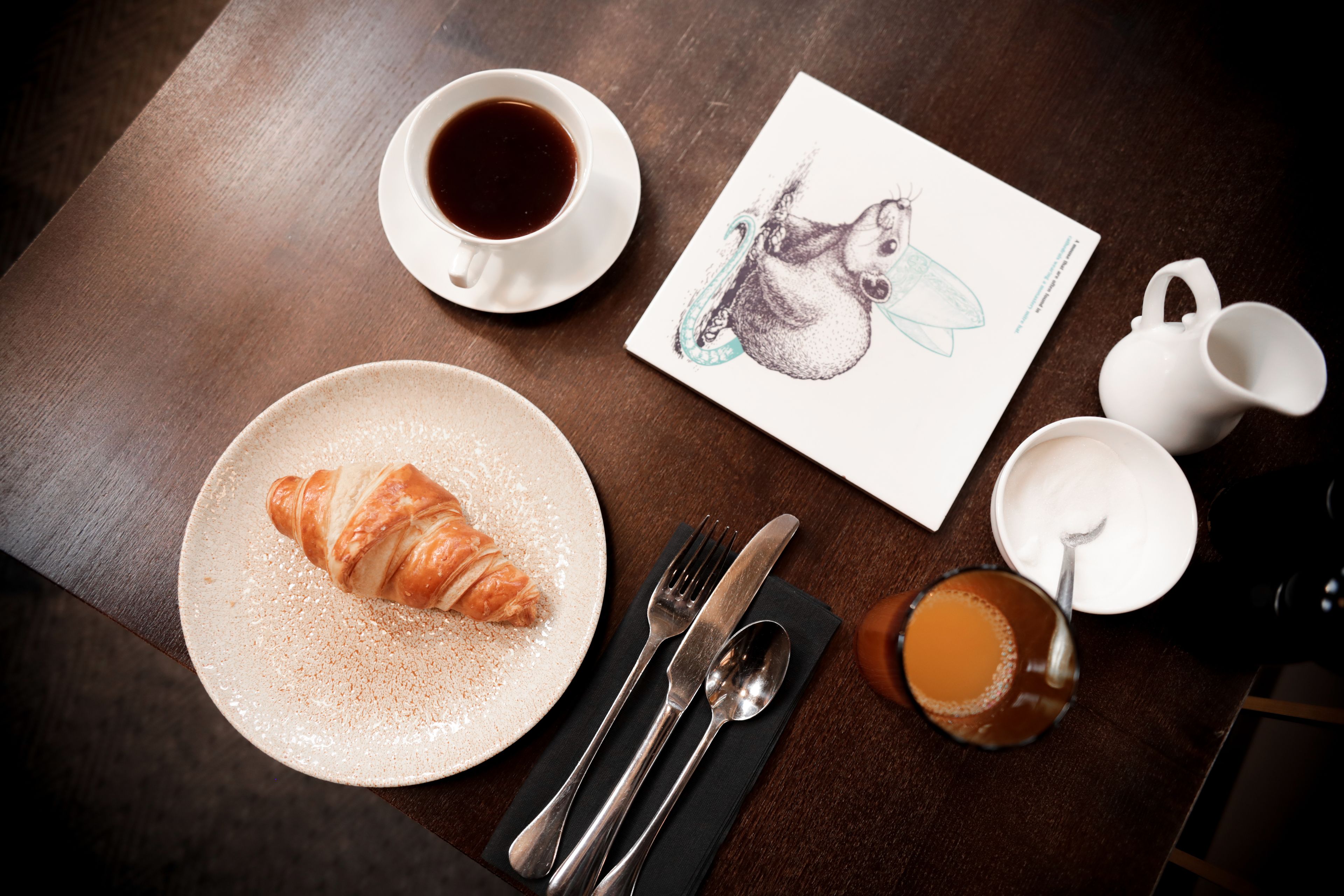 Une table en bois dressée pour le petit-déjeuner, avec un croissant, du café, une tasse de thé et une serviette, ainsi que des couverts.
