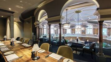 A conference room with a long table set for a meeting, surrounded by chairs, with a view of a lower level through an arched window.