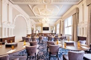 A conference room with multiple tables and chairs, a chandelier, and large windows with curtains.