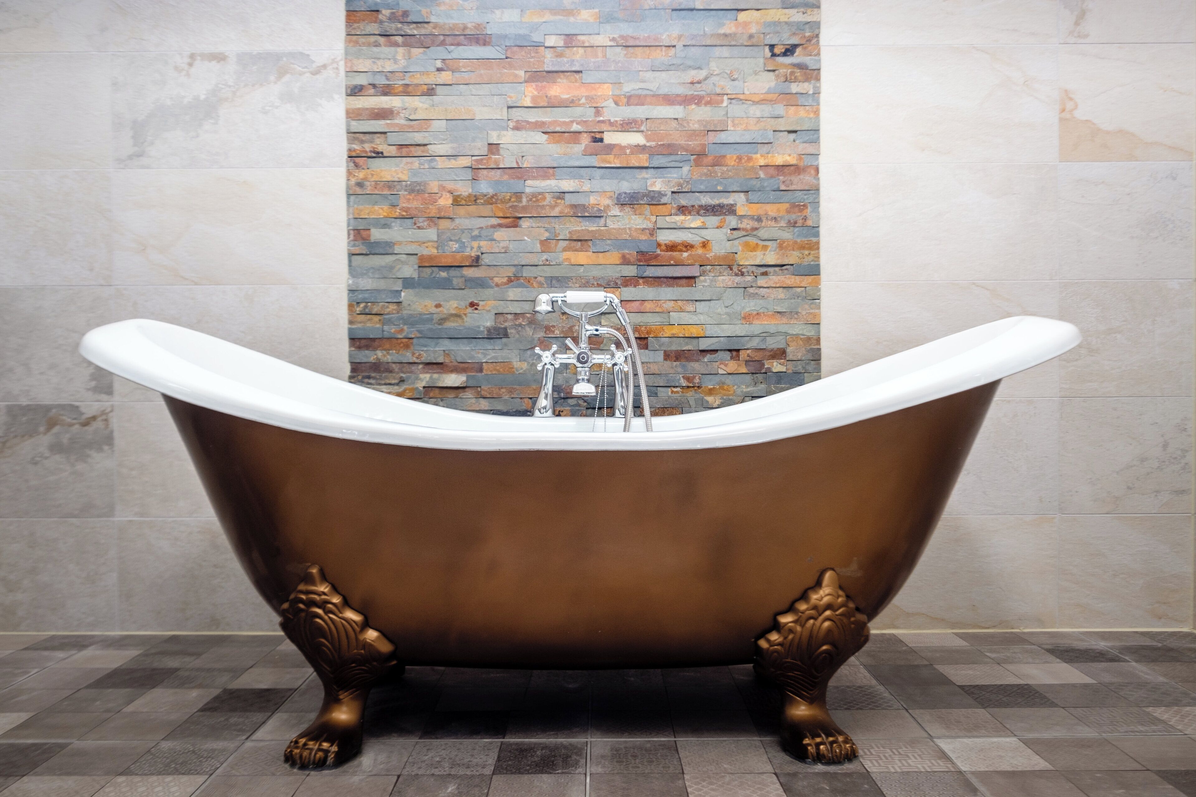Eine freistehende Badewanne auf Füßen, vor einer Wand mit einem steinernen Mosaik-Spritzschutz.
