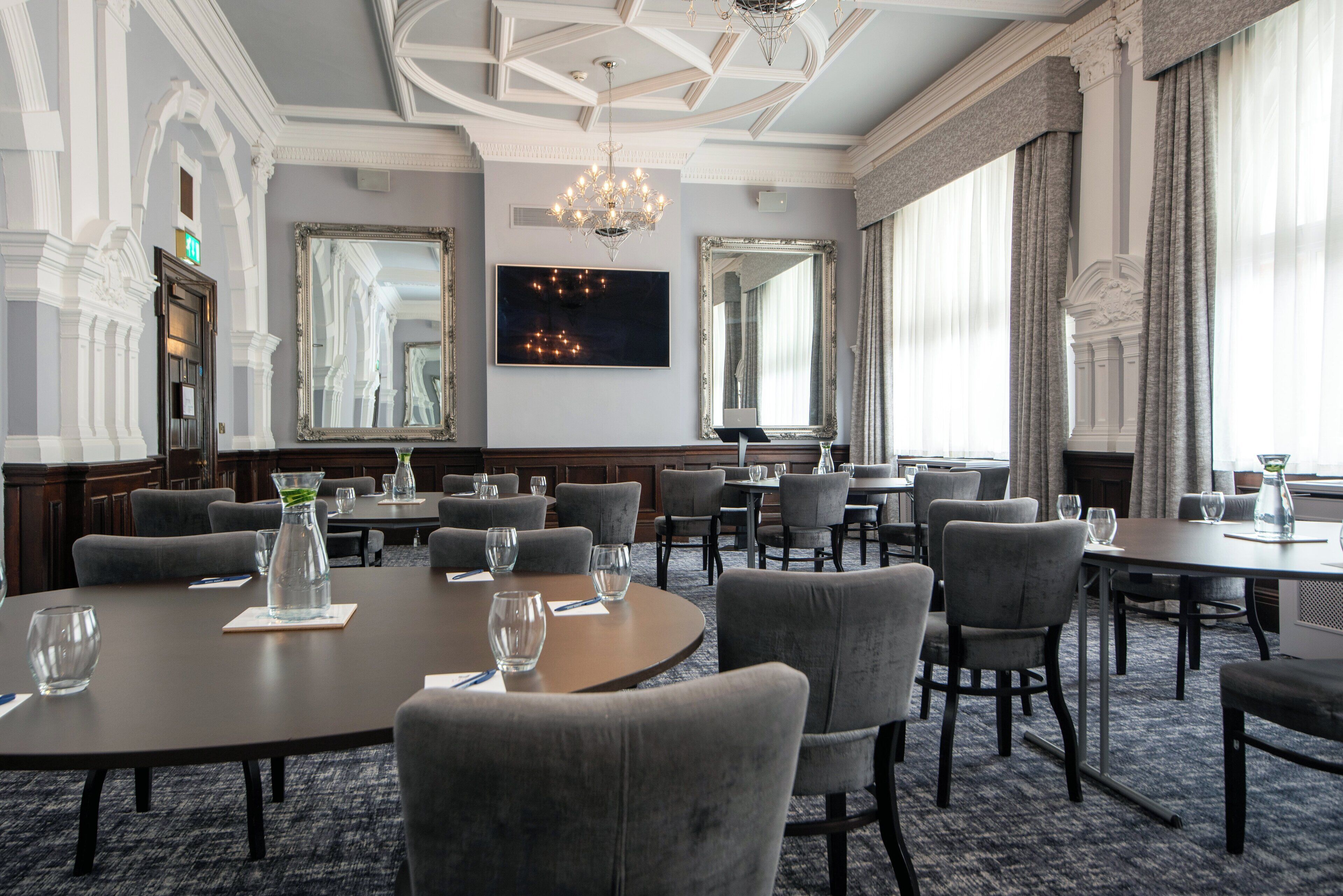 Une salle à manger élégante, avec des tables rondes, des chaises recouvertes d’un tissu gris et des lustres.