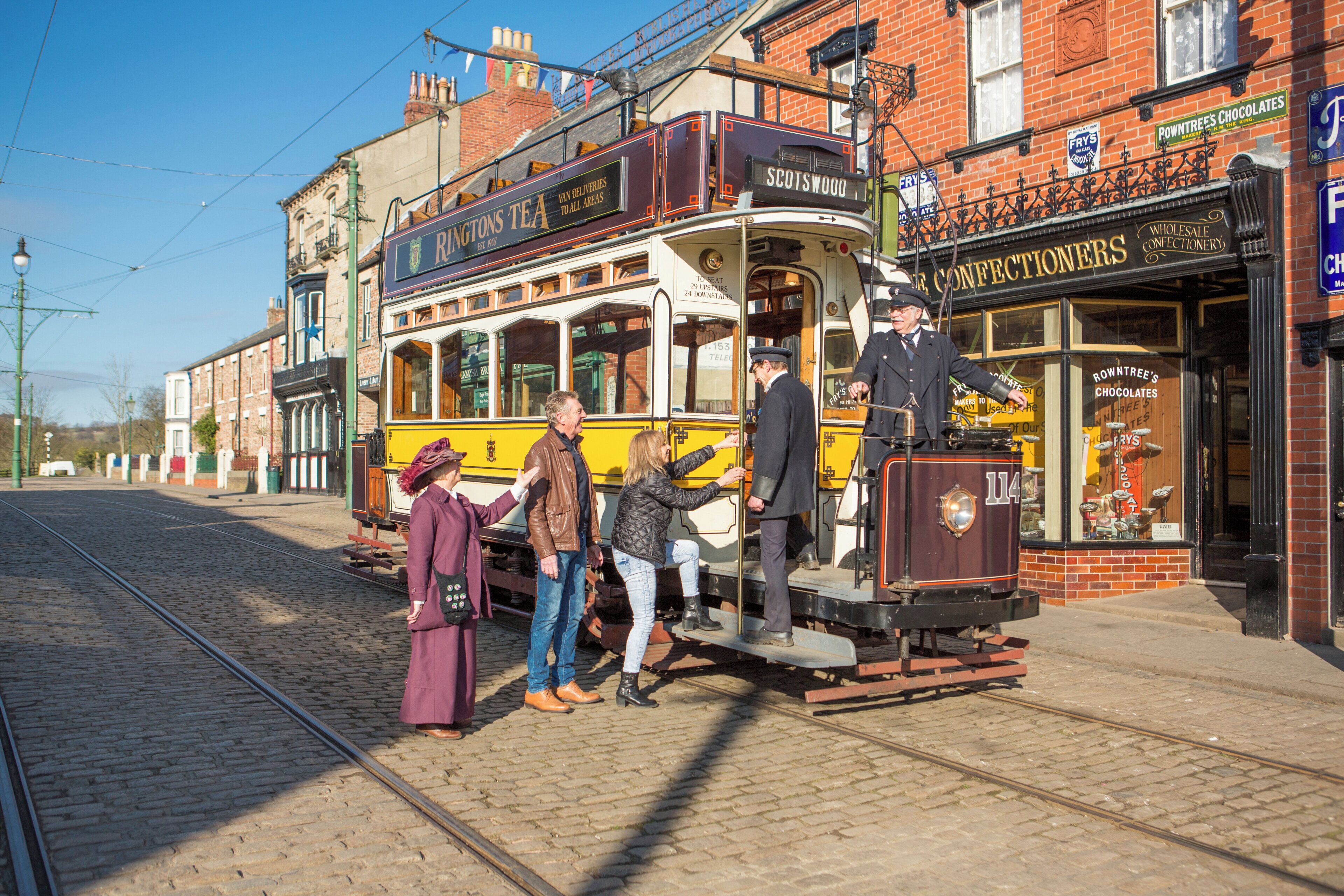 A vintage tram with passengers boarding, set against a backdrop of traditional brick buildings and a clear sky.