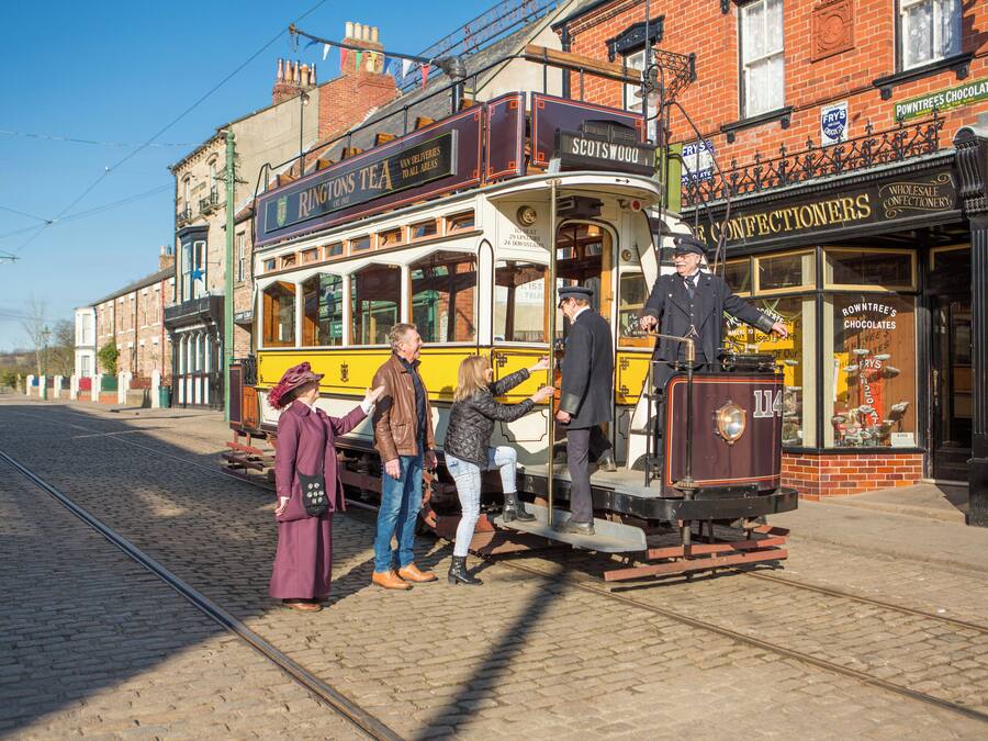 A vintage tram with passengers boarding, set against a backdrop of traditional brick buildings and a clear sky.