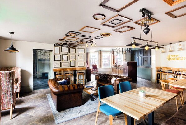 A modern hotel interior with a dining area, a lounge with a leather sofa, and a kitchen visible in the background.