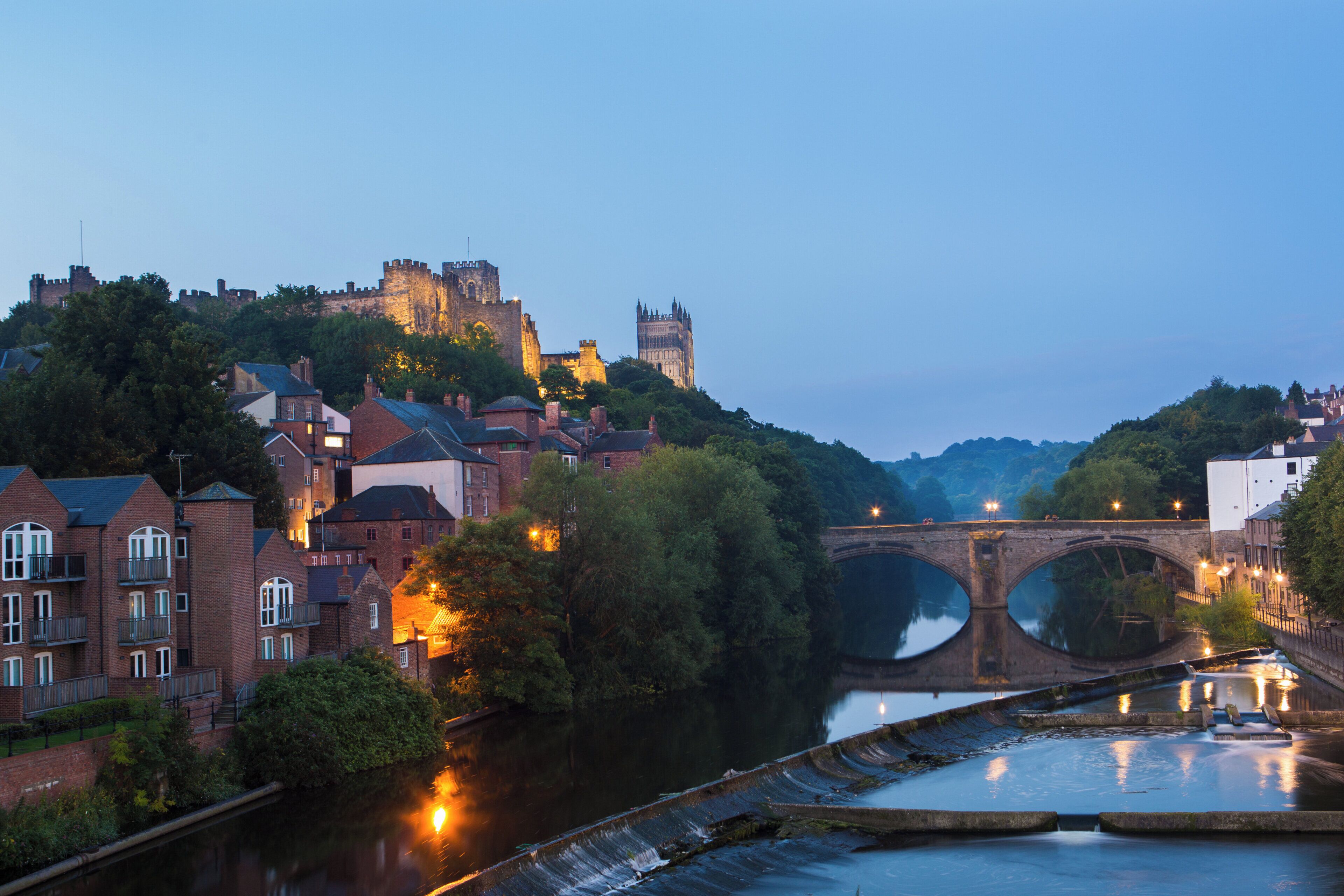 A historic cityscape with a prominent castle on a hill, a bridge over a river, and residential buildings.
