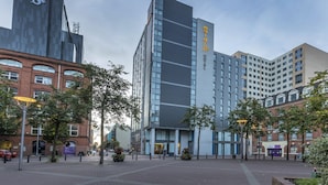 A modern hotel with a prominent logo on the building, surrounded by other urban buildings and trees.