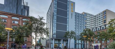 Un hotel moderno con un logotipo destacado en la fachada, rodeado de otros edificios urbanos y árboles.