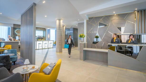 A modern hotel lobby with a reception desk, bar area, and seating with a coffee machine.