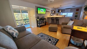 Ein Wohnzimmer mit einer Couch, einem Hocker und einem Couchtisch. An der Wand ist ein Fernseher angebracht, und es gibt eine Küchenzeile mit einer Arbeitsfläche und Barhocker. Eine Glastür öffnet sich zu einem Balkon.