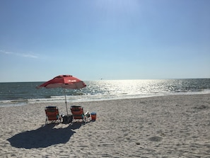 A beach with a red umbrella, two beach chairs, and a cooler.