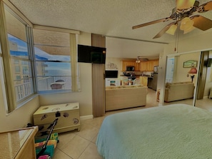 A bedroom with a large bed, a TV, a window with a view, a kitchenette, and a ceiling fan.