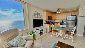 Een woonkamer met een groot raam met uitzicht op zee, een bank met kussens met zeesterren, een televisie en een eethoek met een tafel en stoelen.