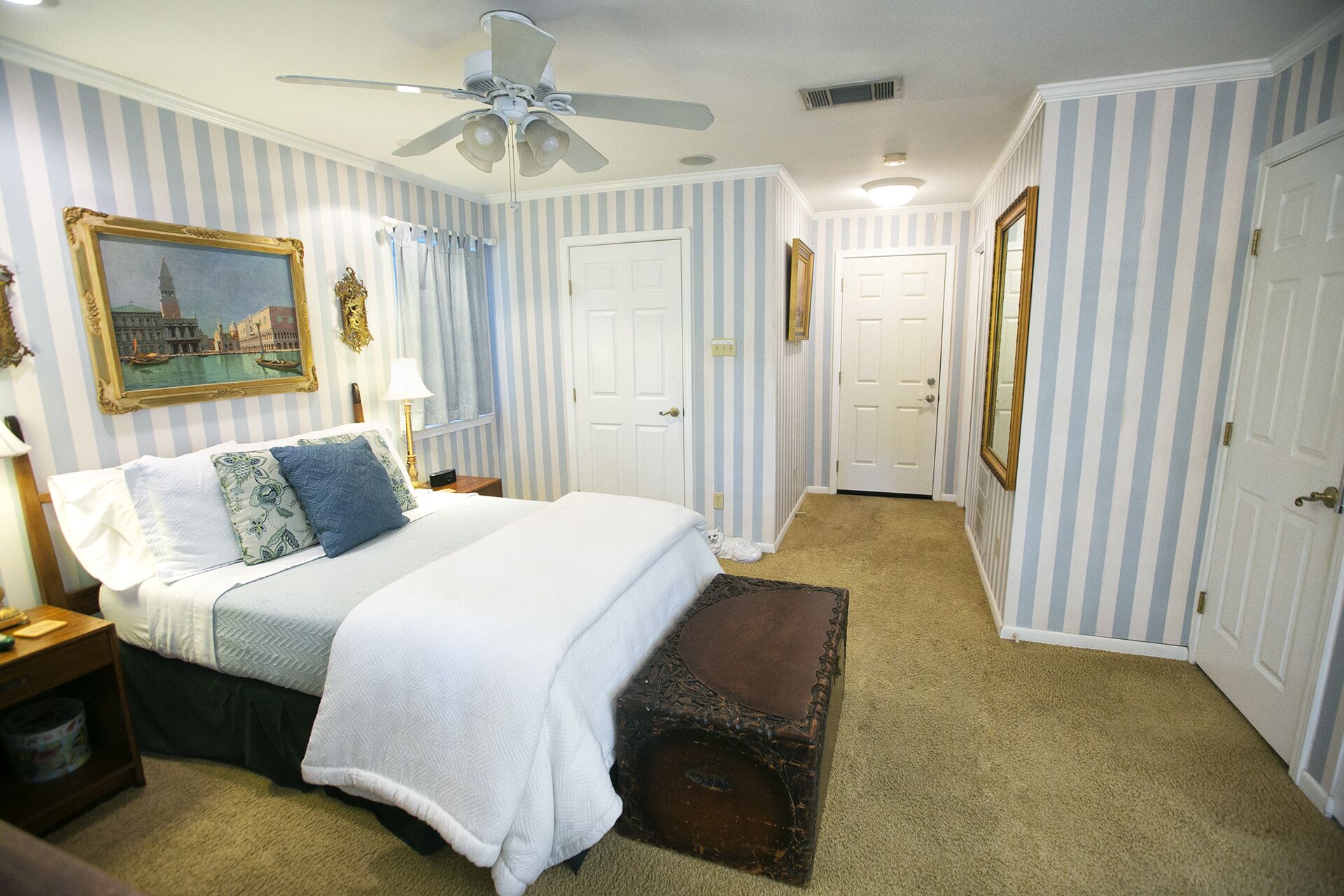 A bedroom with a bed, a ceiling fan, a nightstand, a lamp, a framed picture, and a small dog.
