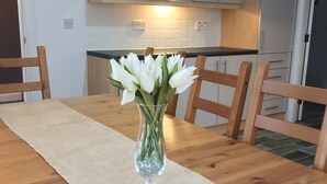 A dining table with a vase of white flowers, wooden chairs, and a kitchen in the background. - The Grange, Portrush. Deluxe holiday home, 8 beds, up to 12 guests, 6 bedrooms.  (Portrush)