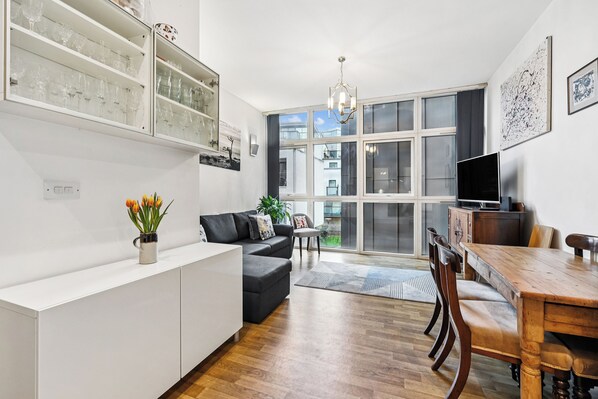 Un salon moderne avec un canapé, une table à manger et un téléviseur. On y trouve des meubles vitrés le long du mur et une grande fenêtre donnant sur des immeubles.