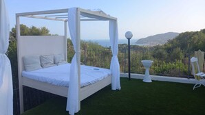 A white four-poster bed with a view of the sea and hills in the background.
